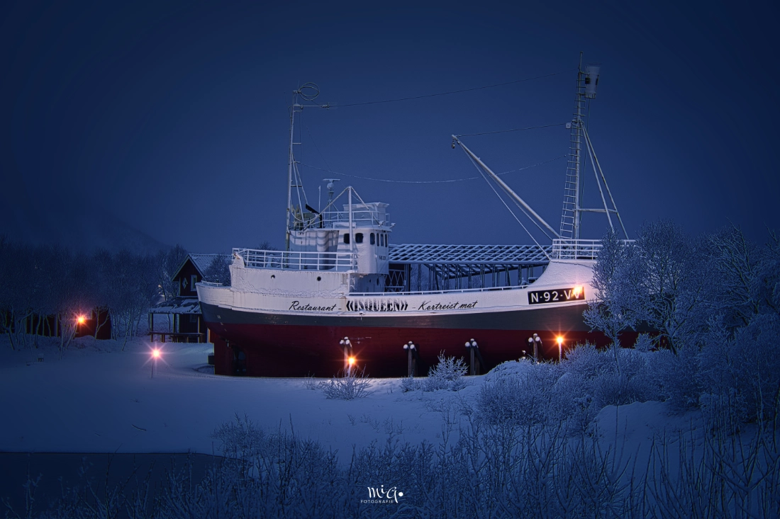 Nordisches Wintermärchen – Das Restaurant-Schiff Ein beleuchtetes Restaurant-Schiff in einer tief verschneiten norwegischen Winterlandschaft während der blauen Stunde