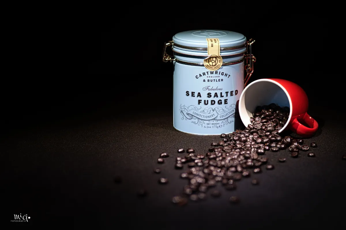 Produktfoto einer hellblauen Dose Sea Salted Fudge neben einer umgekippten roten Tasse mit Kaffeebohnen auf schwarzem Grund