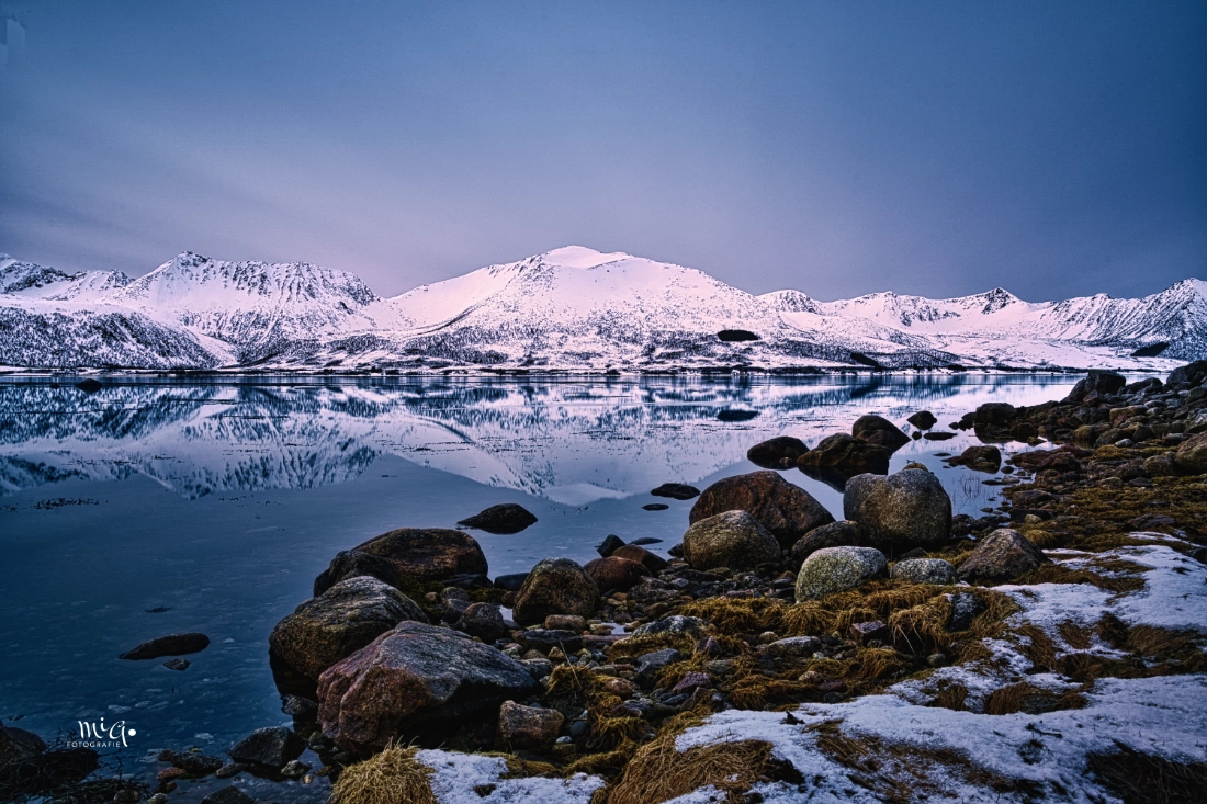 erfekte Spiegelung schneebedeckter Berge in einem eiskalten stillen Fjord der Vesteraalen auf dem Weg nach Andøya in Nordnorwegen mit Granitsteinen im Vordergrund