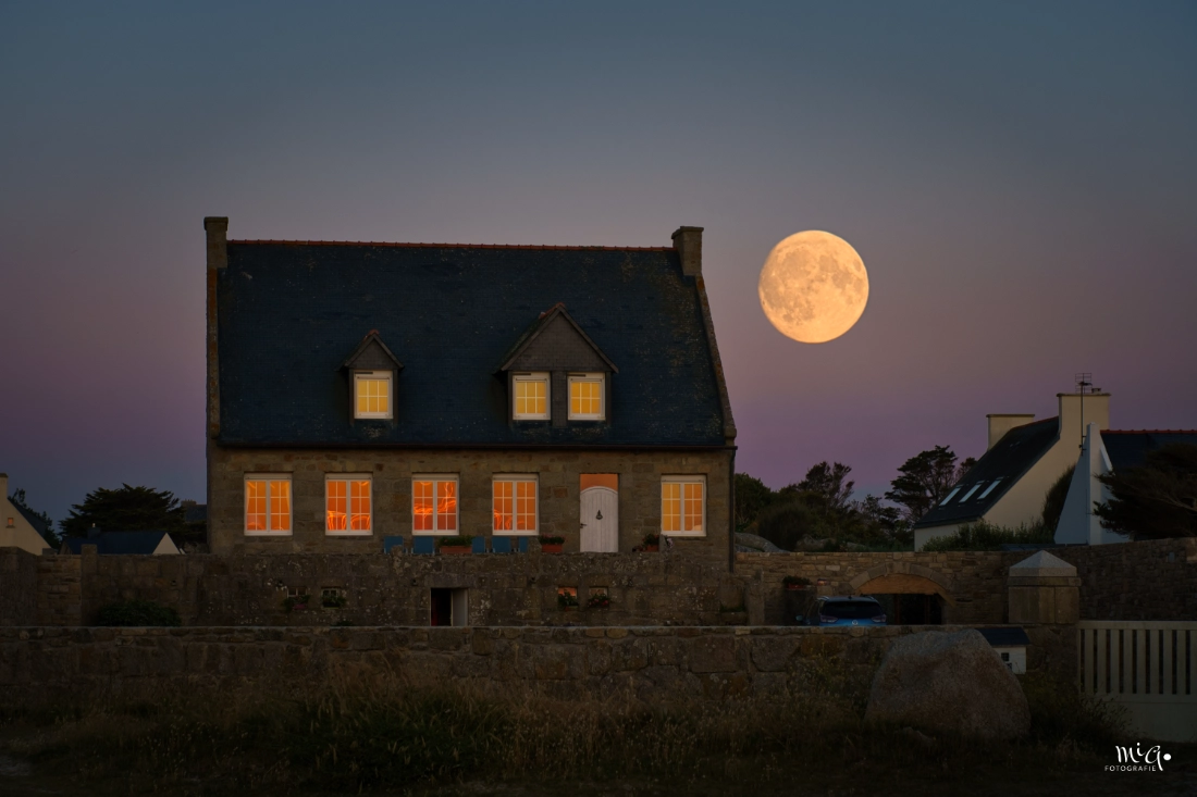 Traditionelles bretonisches Steinhaus mit erleuchteten Fenstern unter einem grossen Vollmond im violetten Abendhimmel der Bretagne
