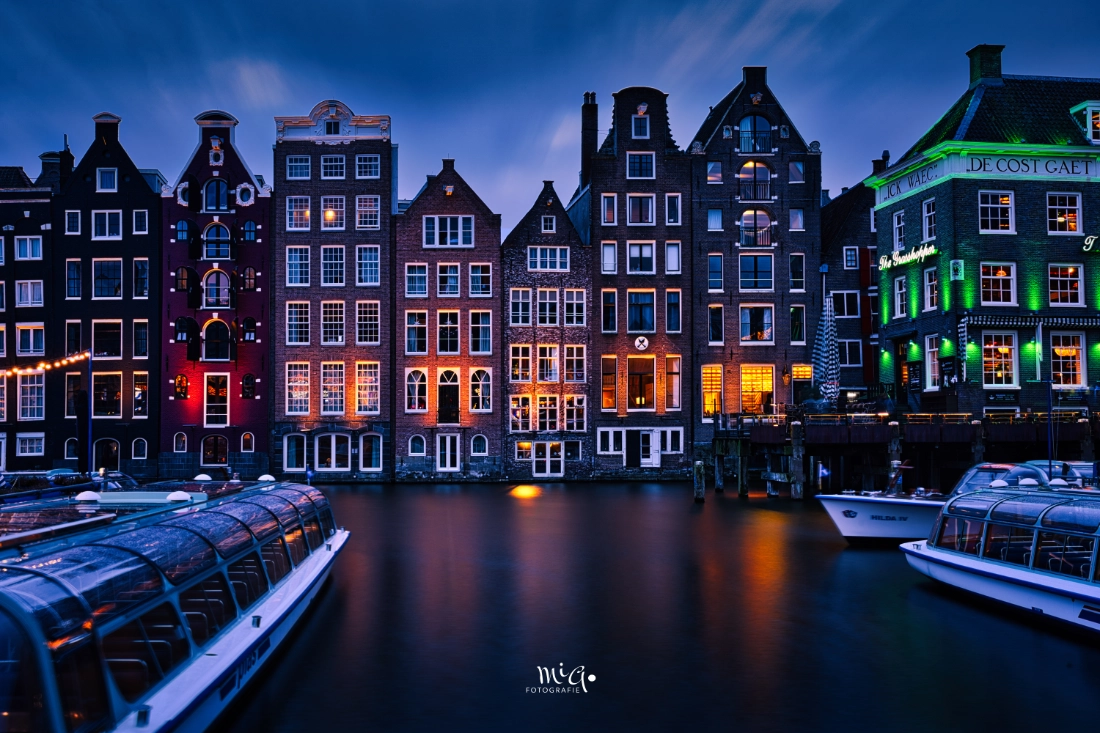 Beleuchtete historische Grachtenhäuser in Amsterdam während der blauen Stunde mit spiegelglattem Wasser durch Langzeitbelichtung und ankernden Rundfahrtbooten.