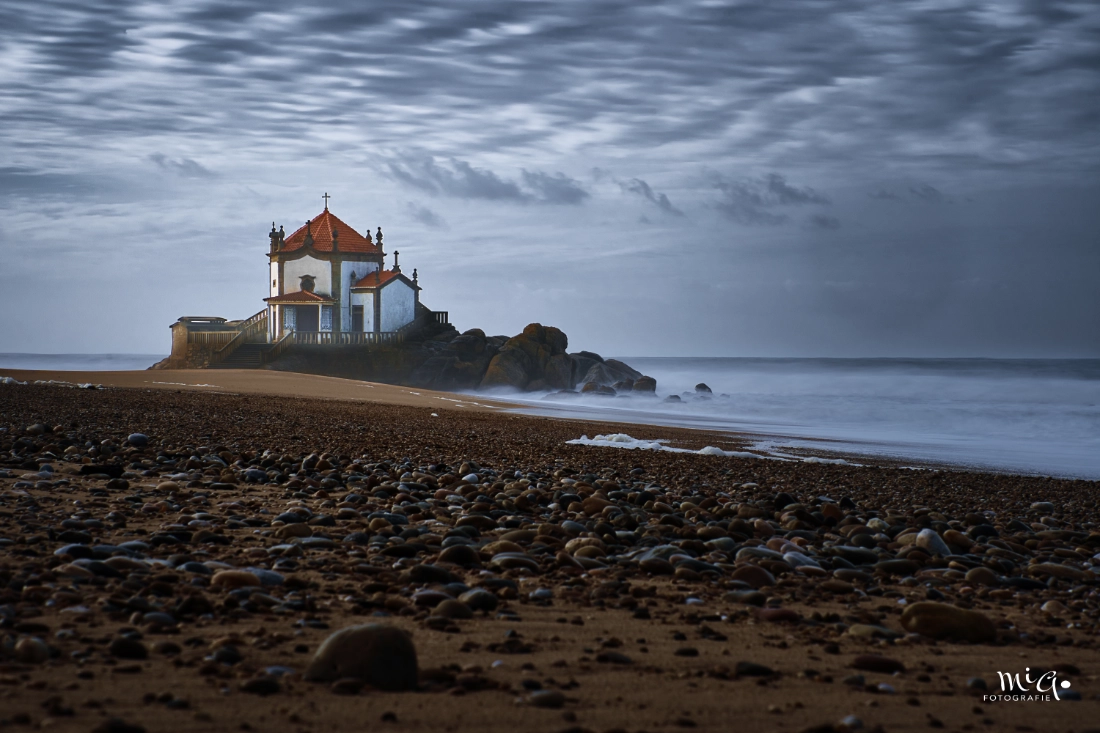 Die Kapelle Capela do Senhor da Pedra an der Küste Portugals, erbaut auf einem Felsen direkt am stürmischen Atlantikstrand bei dramatischem Licht.