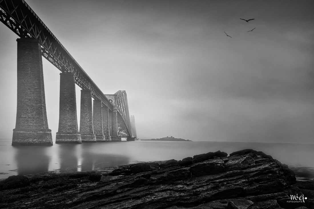 Dramatische Schwarz-Weiß-Aufnahme der Forth Bridge in Schottland mit schroffen Felsen im Vordergrund, Vögeln am Himmel und atmosphärischem Nebel.