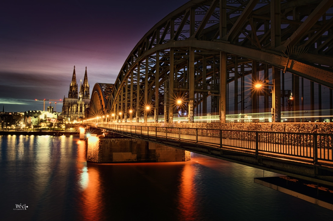 Beleuchteter Kölner Dom und Hohenzollernbrücke bei Nacht mit Lichtspuren eines Zuges, aufgenommen als HDR-Langzeitbelichtung mit Spiegelung im Rhein.