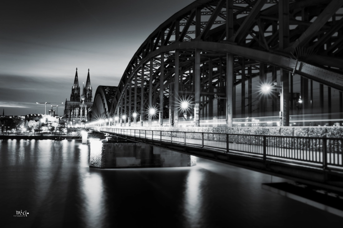Schwarz-Weiß-Aufnahme des Kölner Doms und der Hohenzollernbrücke bei Nacht, mit Lichtspuren vorbeifahrender Züge und Spiegelungen im Rhein.