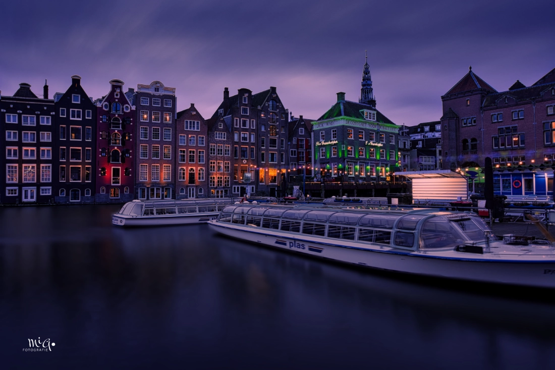 Romantische Abendstimmung an den Amsterdamer Grachten mit beleuchteten historischen Gebäuden und Rundfahrtbooten während der blauen Stunde.