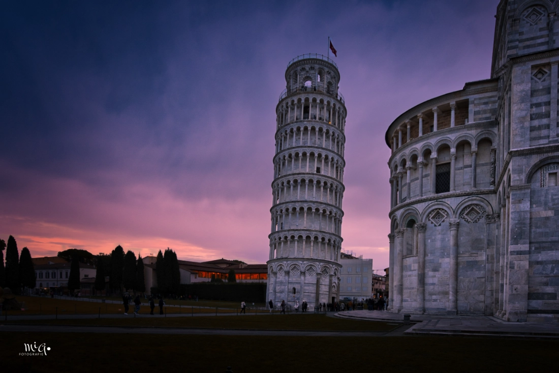 Der Schiefe Turm von Pisa in Italien während der Abenddämmerung, umgeben von historischer Architektur vor einem dramatischen violetten Himmel.