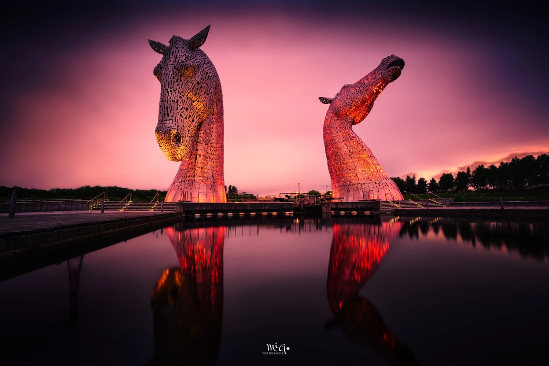 Die Kelpies-Skulpturen in Schottland bei Nacht, beleuchtet und perfekt im ruhigen Wasser gespiegelt, unter einem dramatischen Dämmerungshimmel.