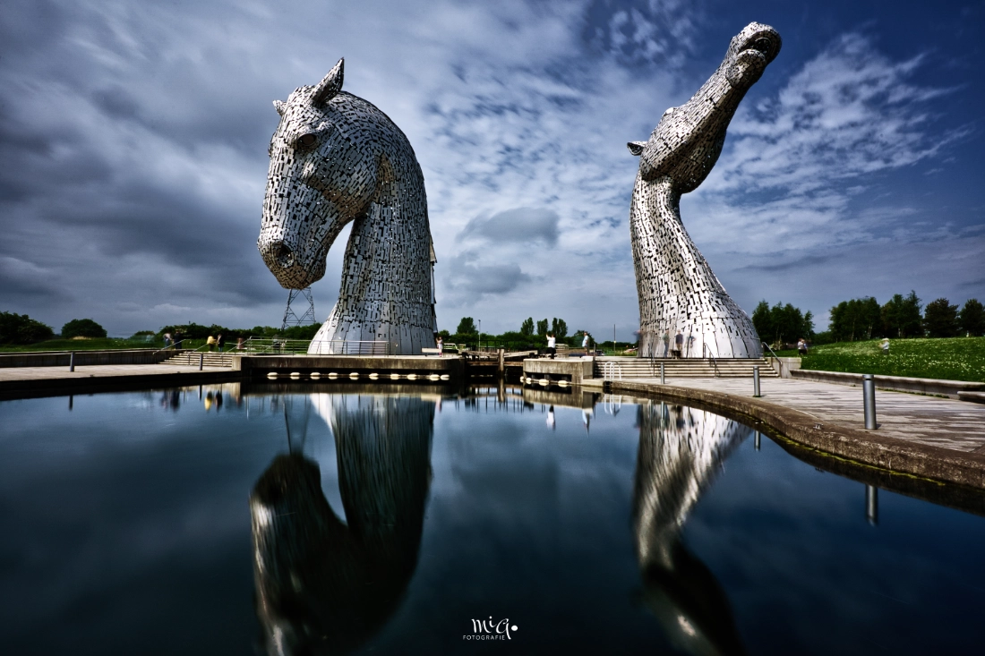 The Kelpies Schottland – Monumentale Stahlskulpturen Die monumentalen Kelpies-Pferdekopfskulpturen aus Stahl in Falkirk, Schottland, unter einem wolkigen Himmel mit Spiegelung im Wasser.